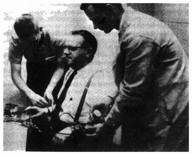 密格兰实验在这张照片上，身穿实验室大褂的实验者和真正的实验对象正把学生（受害者）绑到一张椅子上，并把电极贴到他身上